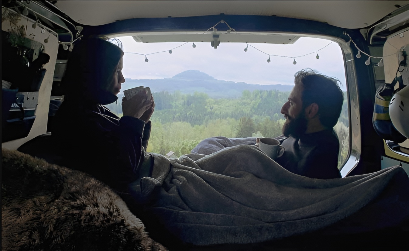 Einblick in den Heckbebreich eines von Vanside ausgebauten Campervans: Ein Pärchen sitzt sich gegenüber bei offener Heckklappe, mit Ausblick auf eine Waldlandschaft und mit Lichterketten und Deko.