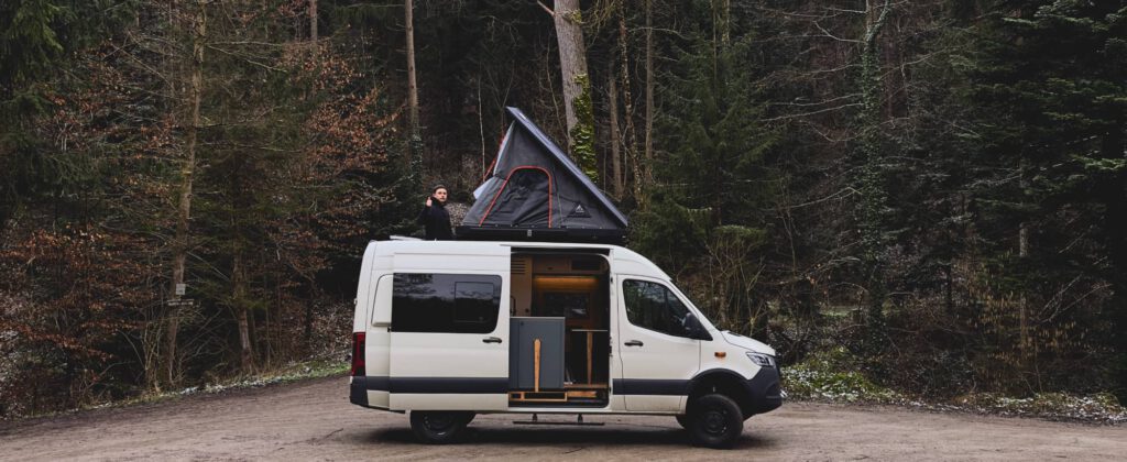 Ein ausgebauter Mercedes Sprinter und das neueste Projekt von Vanside steht auf einem Parkplatz im Wald, das Dachzelt ist ausgeklappt, Geschäftsführer Lukas schaut aus der Dachluke heraus und die Seitentür des Wohnmobils ist geöffnet.