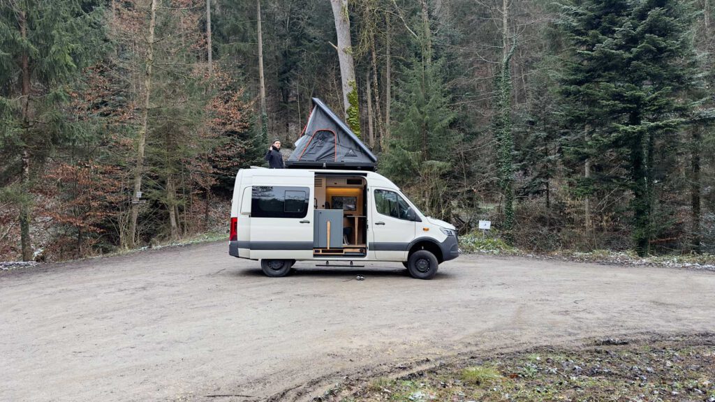 Der neueste Ausbau von Vanside MANNI steht am Waldrand, das Dachzelt ist aufgestellt und Geschäftsführer Lukas schaut heraus. Die Seitentür ist geöffnet und zeigt das Wohnmobil von innen. Vanside hat die ultimative Packliste für eure Campingreisen.