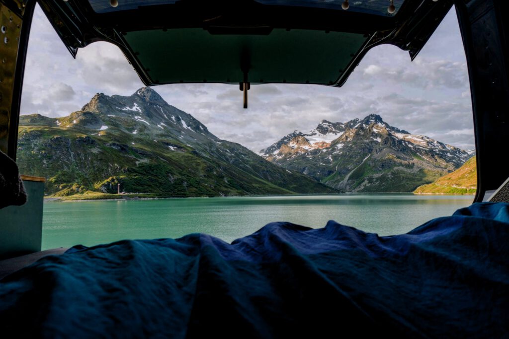 Blick aus dem Kofferraum eines Wohnwagens mit geöffneter Heckklappe und einem Schlafsack, mit Blick auf eine Bergkulisse und einen türkisgrünen See.