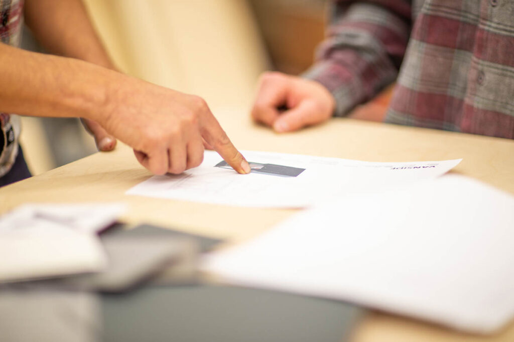 Zwei Personen besprechen einen Bauplan auf einem Tisch. Eine Hand zeigt auf ein Detail auf dem Papier, während die andere Person aufmerksam zuhört. Der Fokus liegt auf den Händen und dem Papier, im Hintergrund sind die Personen leicht verschwommen zu sehen. Der Tisch ist mit verschiedenen Dokumenten und Materialien bedeckt.