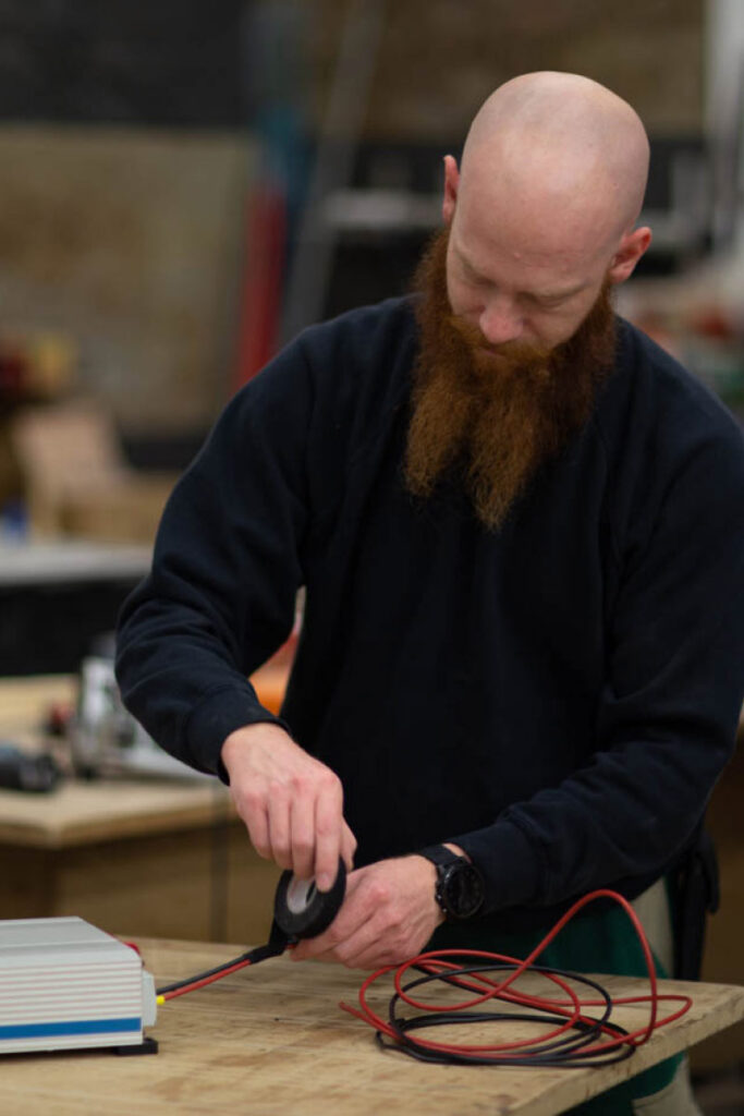 Vanside Elektriker Baste arbeitet in der Werkstatt an einer Batterie und isoliert Kabel neu.