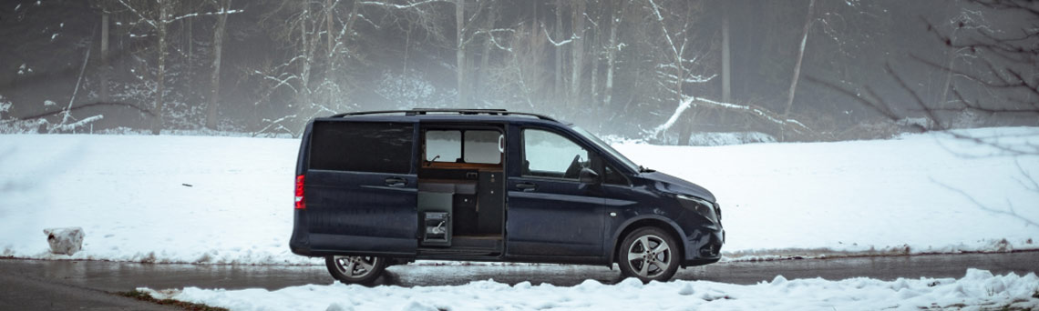 Ein von Vanside ausgebauter Campervan und VW T6 steht auf einem verschneiten Waldrand, die Seitentür ist geöffnet.