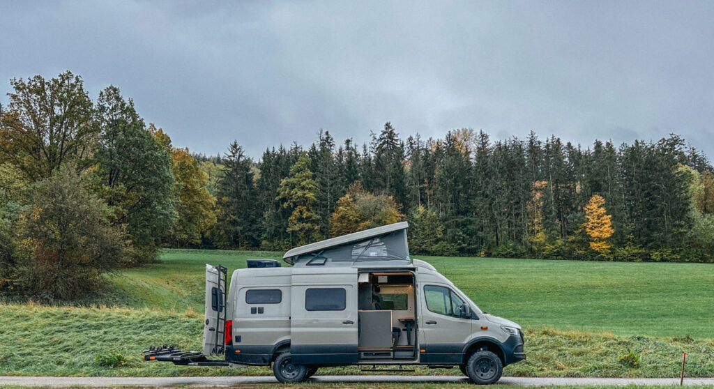 Der neue Vanside Camper Ausbau LEO, ein hellgrauer Mercedes-Benz Sprinter, steht vor einem Waldstück, die Seitentür und Hintertür sind geöffnet, das Dachzelt ist geöffnet.