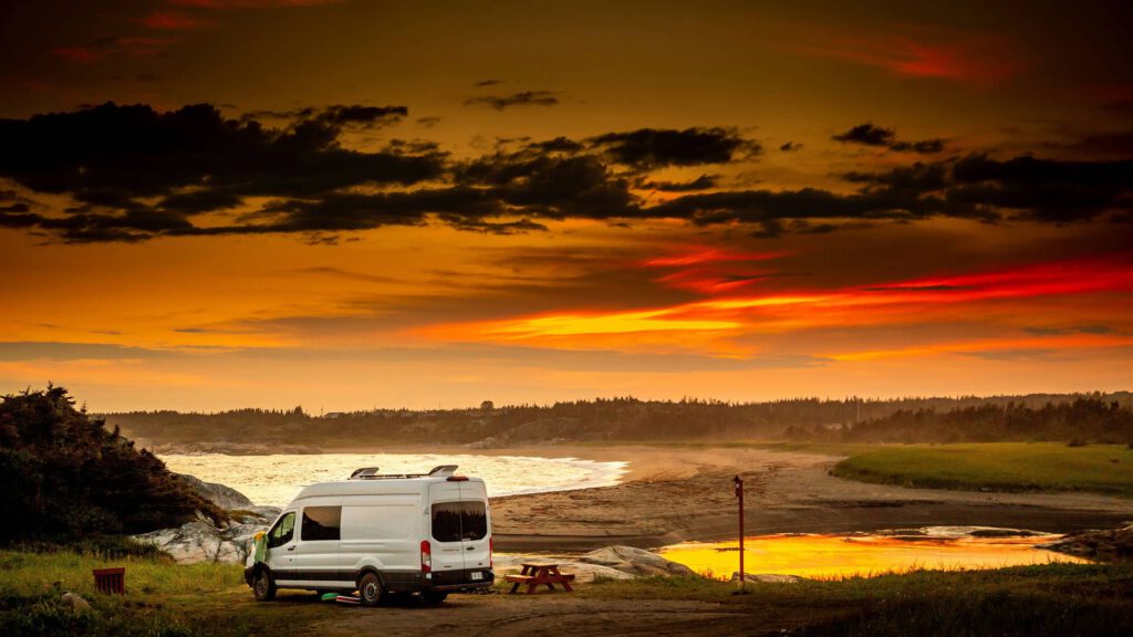 Ein weißes Wohnmobil steht auf einer Wiese am Rande eines Strandes bei Sonnenuntergang. Der Himmel ist in warmen Gelb- und Orangetönen gefärbt, mit dunklen Wolken, die über den Horizont ziehen. Im Hintergrund sind Bäume und die Küstenlinie zu sehen, die sich entlang des Strandes erstrecken.
