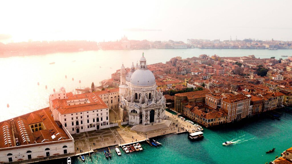 Luftaufnahme von Venedig, Italien, mit der Kirche Santa Maria della Salute im Vordergrund, umgeben von roten Dächern und Wasserstraßen. Im Hintergrund ist die Lagune von Venedig mit einigen Booten zu sehen. Der Himmel ist hell und sonnig.