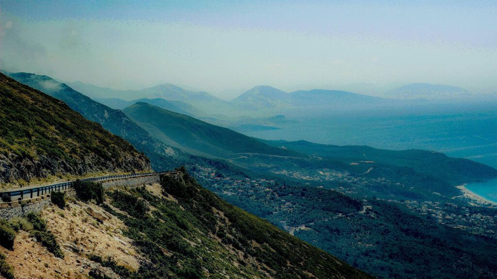 Eine gewundene Küstenstraße, die sich entlang eines Berges erstreckt, mit Blick auf das tiefblaue Meer und die umliegenden Hügel im Hintergrund.